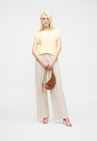 Γυναίκα που φοράει ανοιχτόκίτρινη μπλούζα με λεπτομέρειες ανοιχτών ματιών και μπεζ φαρδύ παντελόνι, κρατώντας καφέ δερμάτινη τσάντα, στέκεται σε λευκό φόντο.