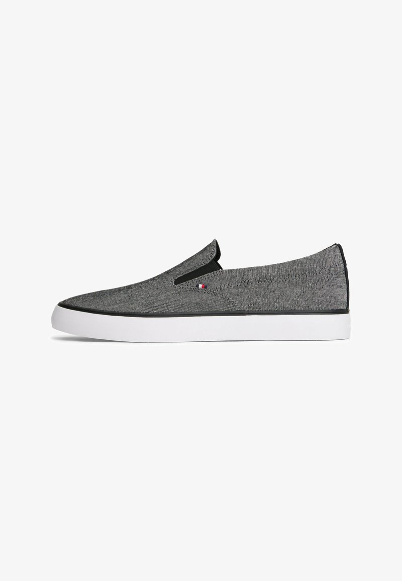 Zapatilla deportiva gris sin cordones con suela de goma blanca y un pequeño logo rojo, blanco y azul en el lateral, sobre un fondo blanco.