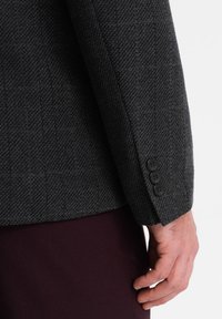 Grå herringbone-jacka i blandmaterial med tre svarta knappar på ärmen, bärs över ett bourgogne-färgat tyg. Strukturerad axel och texturerat tyg.
