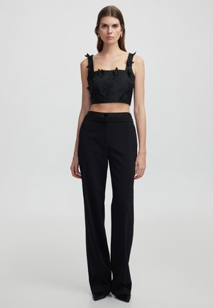 Zwarte crop top met bloemtextuur en details van bandjes, gecombineerd met hoge zwarte broeken met een rechte pijp en knoopsluiting.