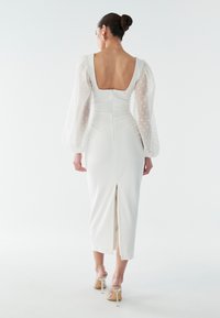 Vit figursydd klänning med genomskinliga puffärmar med prickar, fyrkantig rygg och slits bak, bärs med klara högklackade sandaler.