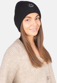 Bonnet noir en tricot avec une broderie de visage souriant à l'avant, porté avec un cardigan en tricot beige clair avec des boutons dorés.