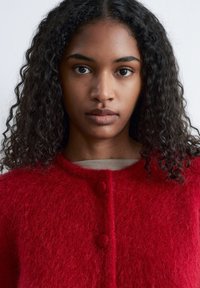 Roter, flauschiger Wollstrickjacke mit runden Knöpfen und hohem Kragen. Weiche Textur und schlichtes Design, getragen über einem hellfarbenen Hemd.