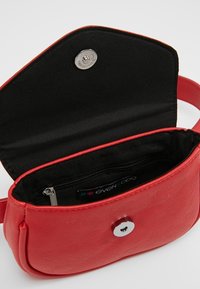 Punainen nahkainen olkalaukku pyöreällä muotoilulla, napilla suljettava ja musta sisustus, jossa on vetoketjullinen tasku ja brändilogo.