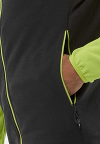 Black fleece jacket with a textured surface, featuring a green zipper accent and cuff. Hand in pocket highlights practical design.