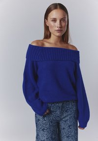 Koboltblå off-shoulder tröja med ribbad halsringning. Mjuk, texturerad tyg och lösa ärmar. I kombination med mönstrade blommiga jeans.