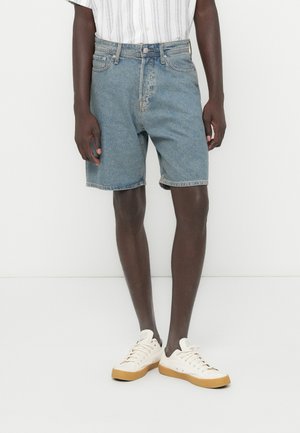 Shorts in denim blu con una vestibilità comoda, caratterizzati da un design con cinque tasche, orli risvoltati e una finitura lavata chiara, abbinati a scarpe da ginnastica bianche.