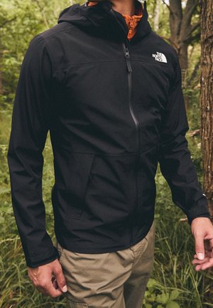 Osoba ubrana w czarną kurtkę The North Face z kapturem i beżowe spodnie, stojąca na zewnątrz obok drzew i zieleni.