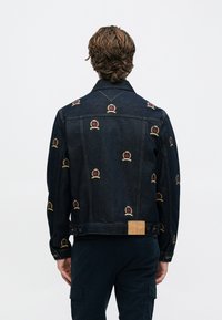 Σκούρο μπουφάν από τζιν με κεντημένα μοτίβα κορώνας σε κόκκινο και χρυσό, με κουμπιά μπροστά και δερμάτινο ταμπελάκι της μάρκας στη μέση.