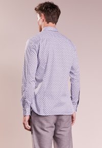 Man som visas bakifrån, klädd i en vit långärmad skjorta med ett litet mörkt geometriskt mönster och grå byxor mot en beige bakgrund.