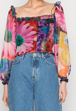 Mujer con una blusa transparente de manga larga con estampado floral colorido, combinada con vaqueros de mezclilla azul de cintura alta.