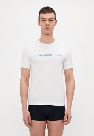 T-shirt bianca a maniche corte realizzata in morbido tessuto, con logo "EMPORIO ARMANI" in blu. Il modello indossa biancheria intima nera. Design semplice, scollo rotondo.