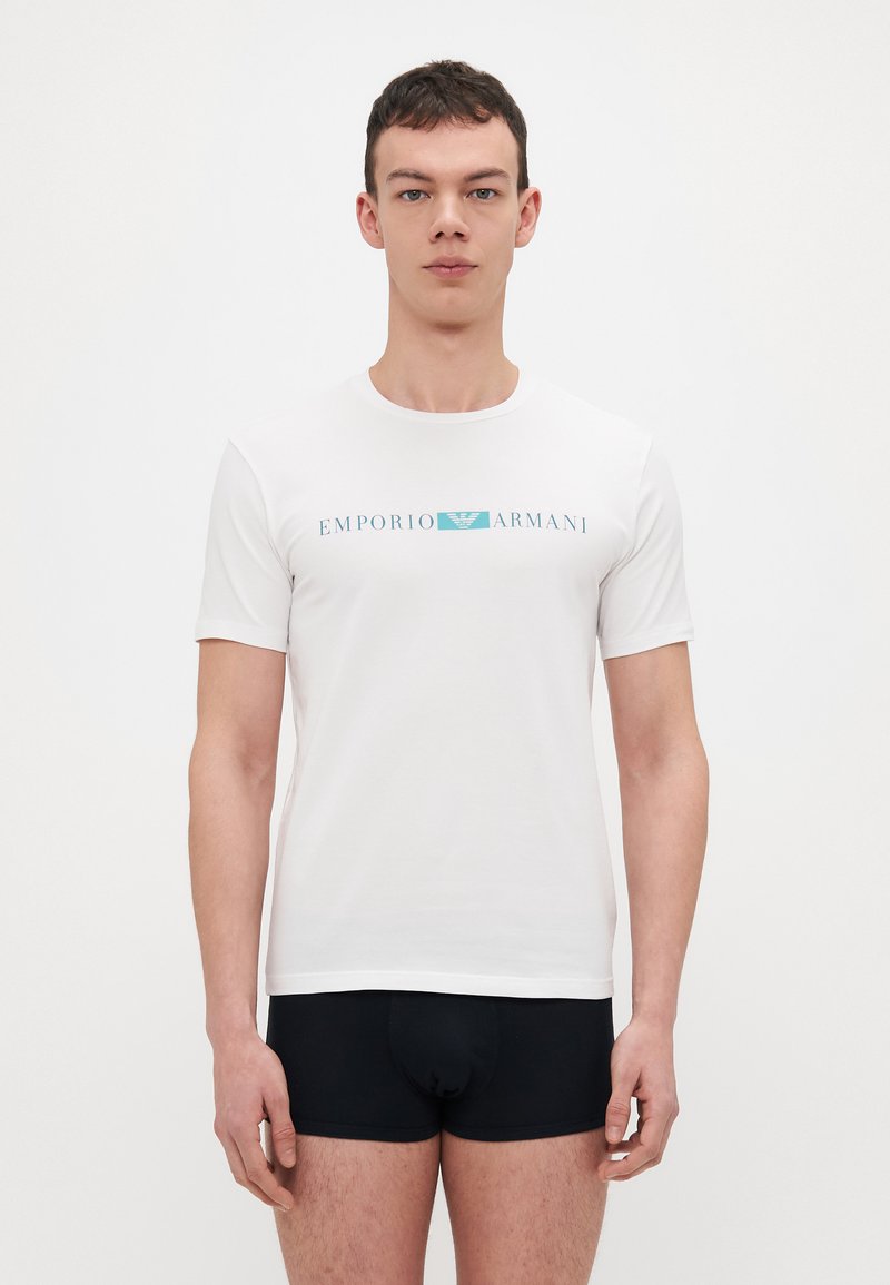 Balta trumpomis rankovėmis marškinėliai pagaminti iš minkštos medžiagos, su mėlynu „EMPORIO ARMANI“ logotipu. Modelis dėvi juodus apatinius. Paprasto dizaino, apvaliu iškirpimu.