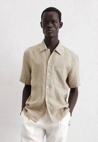 Camicetta a maniche corte a righe beige realizzata in tessuto leggero, con taschino e bottoni, abbinata a pantaloni in lino bianchi.