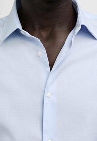 Camicia azzurro chiaro con colletto corto, realizzata in un tessuto testurizzato e con cinque bottoni visibili sul davanti. Design liscio e strutturato.