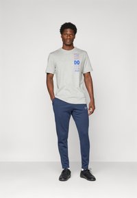Mies, der ein graues "Just Do It." T-Shirt, eine marineblaue Nike-Sweatpants und schwarze Sneakers trägt, steht vor einem einfarbigen hellen Hintergrund.