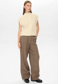 Gebreide, mouwloze lichtbeige top met een hoge hals en kabelpatroon, gecombineerd met losse, bruine, op maat gemaakte broeken en zwarte plateauschoenen.