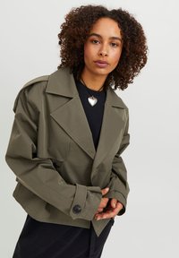 Lyhyt oliivinvihreä trench-coat, jossa on suuret huippukäänteet, kaksirivinen nappikiinnitys ja teksturoitu kangas. Yhdistetty mustan paidan kanssa.