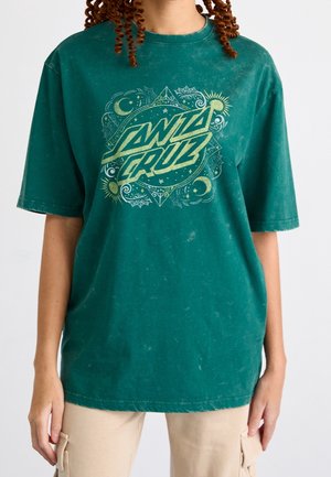 T-shirt oversize turquoise avec le logo "Santa Cruz" entouré de motifs célestes, porté avec un pantalon cargo beige.