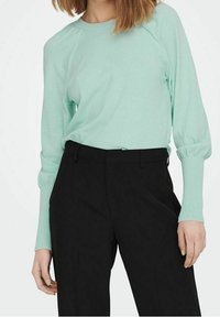 Pull vert menthe à manches longues avec un col rond et des manches bouffantes, associé à un pantalon noir sur mesure de coupe classique.