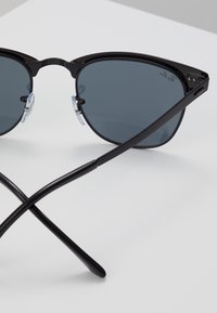 Occhiali da sole cat-eye neri con lenti grigie, montatura nera sottile e padiglioni trasparenti. Le aste sono eleganti con un design angolare, incrociate in primo piano.