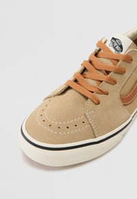 Beige nubuk sneaker barna díszítésekkel, narancs fűzőkkel, perforációkkal, fehér varrással és fekete csíkkal ellátott gumitalppal.