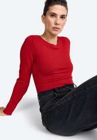 Rød ribbet langærmet top med rund halsudskæring, parret med højtaljede sorte jeans. Stoffet ser glat ud med en tætsiddende silhuet.