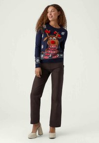 Marineblauer Pullover mit einem Rentierdesign, roter Nase und Schal, Schneeflöckchen und gemustertem Saum. Kombiniert mit braunen Hosen und cremefarbenen Absatzschuhen.