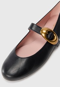 Chaussure Mary Jane en cuir noir avec une large boucle dorée et une semelle intérieure rose douce sur fond blanc.