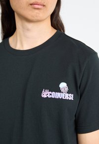Sort bomulds T-shirt med et grafik af en meduse og koraller i pink og blå, med "Converse" tekst i boblebogstaver på brystet.