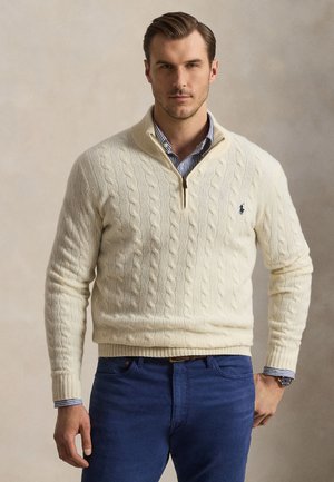 Man draagt een crème kabelgebreide trui met kwartrits over een blauw gestreept overhemd en een blauwe broek, staand tegen een effen beige achtergrond.