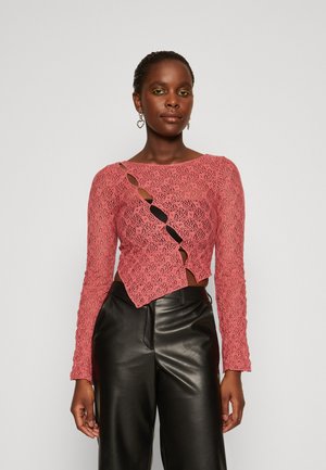 Pink lace long-sleeve top featuring cut-out detailing and a curved hem, paired with black leather pants. Textured design with intricate patterns.
