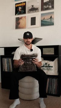Uma pessoa segura um saco de plástico transparente que contém uma camisa marrom dobrada. O fundo apresenta discos de vinil e uma coluna de som.