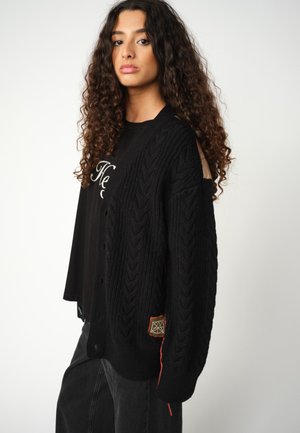 Donna con lunghi capelli ricci che indossa un cardigan di lana nera a trecce sopra una maglietta nera e pantaloni scuri, in piedi davanti a uno sfondo bianco semplice.
