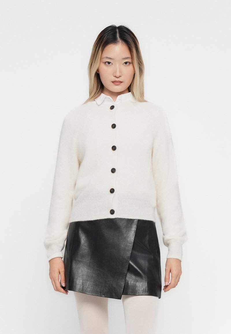 Vrouw in een witte knooptrui over een blouse met kraag en een zwarte leren minirok, staande tegen een effen witte achtergrond.