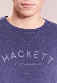 Parte inferior de la cara y cuello de una persona que lleva una sudadera de cuello redondo azul con el texto "HACKETT LONDON·ENGLAND" en el pecho.