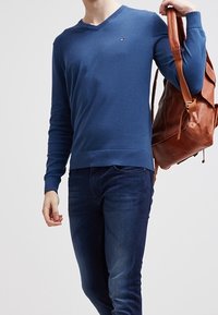 Niebieski bawełniany sweter z dekoltem w serek, noszony z ciemnymi dżinsami. Na jednym ramieniu wisi brązowy skórzany plecak, który nadaje kontrast całości.