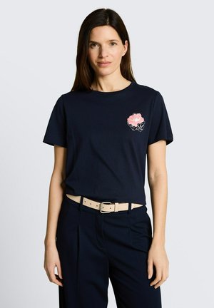 Kobieta o długich brązowych włosach, ubrana w granatowy t-shirt z różowym wzorem kwiatu na piersi oraz beżowy pasek i granatowe spodnie.