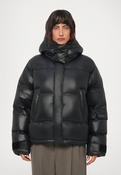 Eine Person trägt eine schwarze Steppjacke mit hohem, gepolstertem Kragen, Klettverschlussriemen an den Ärmeln und grauen Hosen. Sie steht frontal vor einem weißen Hintergrund.