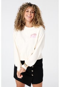 Crème lange mouw top met de tekst "QUEEN OF HEARTS" in het roze, gecombineerd met een zwarte rok met hartvormige knopen. Zacht materiaal, ontspannen pasvorm.