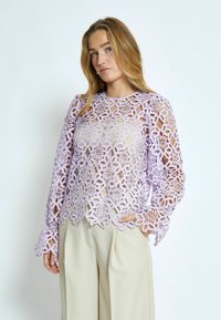 Blusa di pizzo lavanda con intricate incisioni, maniche lunghe con polsini a campana e scollatura rotonda. Abbinata a pantaloni beige.