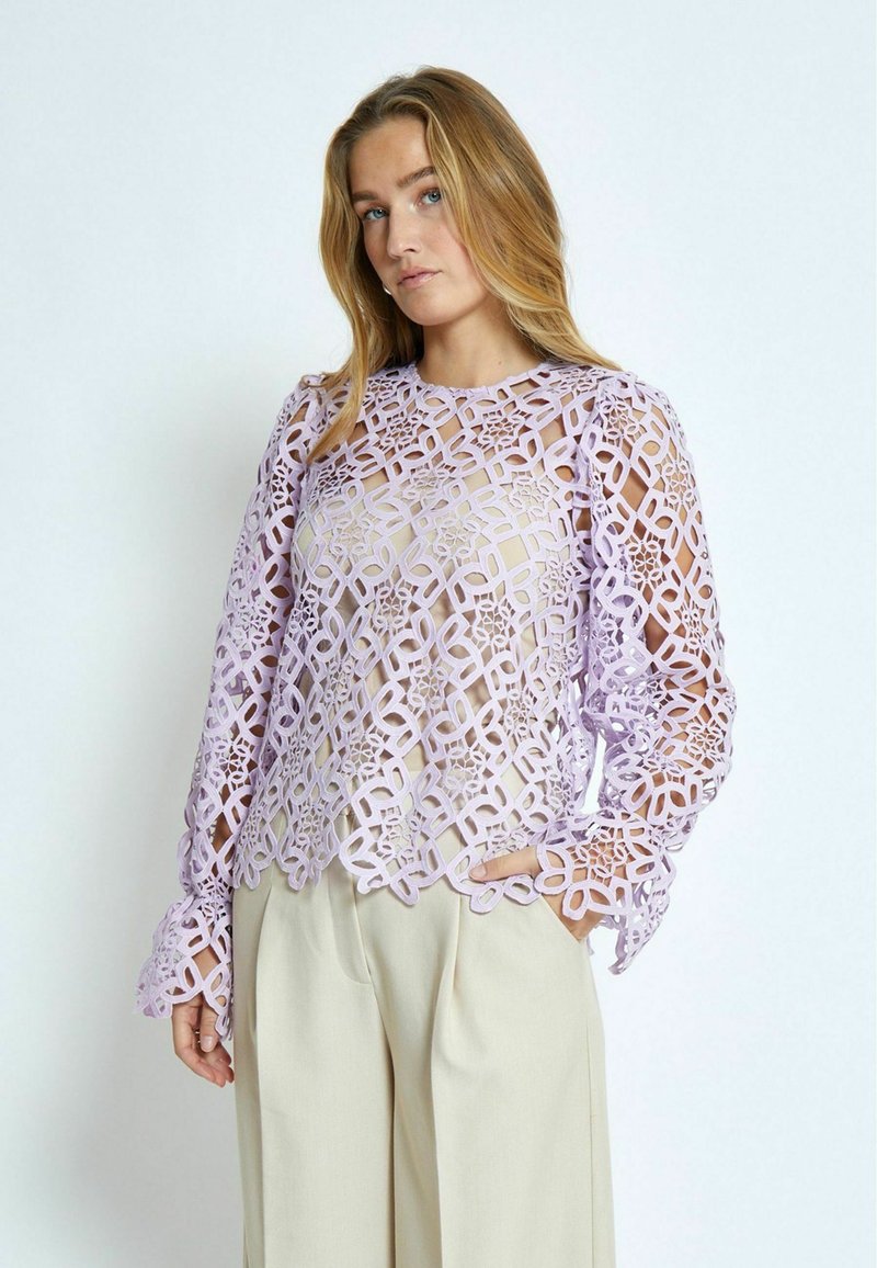 Blusa di pizzo lavanda con intricate incisioni, maniche lunghe con polsini a campana e scollatura rotonda. Abbinata a pantaloni beige.