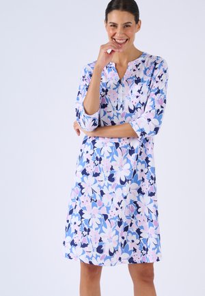 Femme souriante portant une robe mi-longue à motifs floraux roses, bleus et blancs, debout avec un bras croisé et la main près du visage.