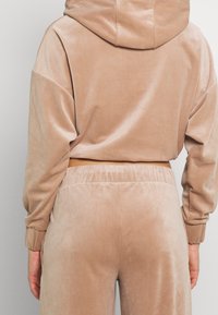 Tuta in velluto beige con top con cappuccio, spalle scese e pantaloni con vita elastica, che evidenzia una texture morbida e liscia.