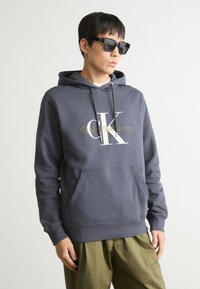 Siva bombažna kapuca s sprednjim žepom, ki vsebuje belo in zlato "Calvin Klein Jeans" logotip. V kombinaciji z olivno zelenimi hlačami in sončnimi očali.