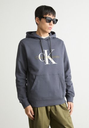 Grå bomullshoodie med frontlomme, med en hvit og gull "Calvin Klein Jeans" logo. Sammen med olivengrønne bukser og solbriller.
