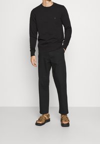 Felpa nera in cotone con scollo rotondo, abbinata a pantaloni neri a gamba larga e sandali marroni con cinturini. Design minimalista con marchio discreto.