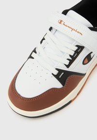 Λευκό, καφέ και μαύρο αθλητικό παπούτσι με λευκά κορδόνια και λουράκι με την ένδειξη "Champion" σε λευκό φόντο.