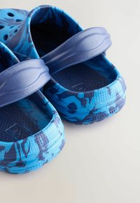 Blå mönstrade skor med en slip-on-design, som har en tjock sula och ett bälte på baksidan för stöd. Texturerad inre yta för grepp.