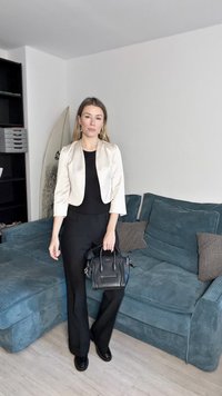 Černé kalhoty a top doplněné krémovým sakem. Modelka drží černou kabelku a stojí na texturované modré pohovce v moderním interiéru.